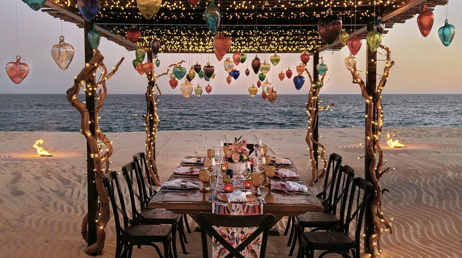 A dinner table on the beach at dusk under strings of lights and glass hearts
