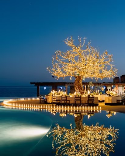 A lit tree over a dinner table beside the pool at night