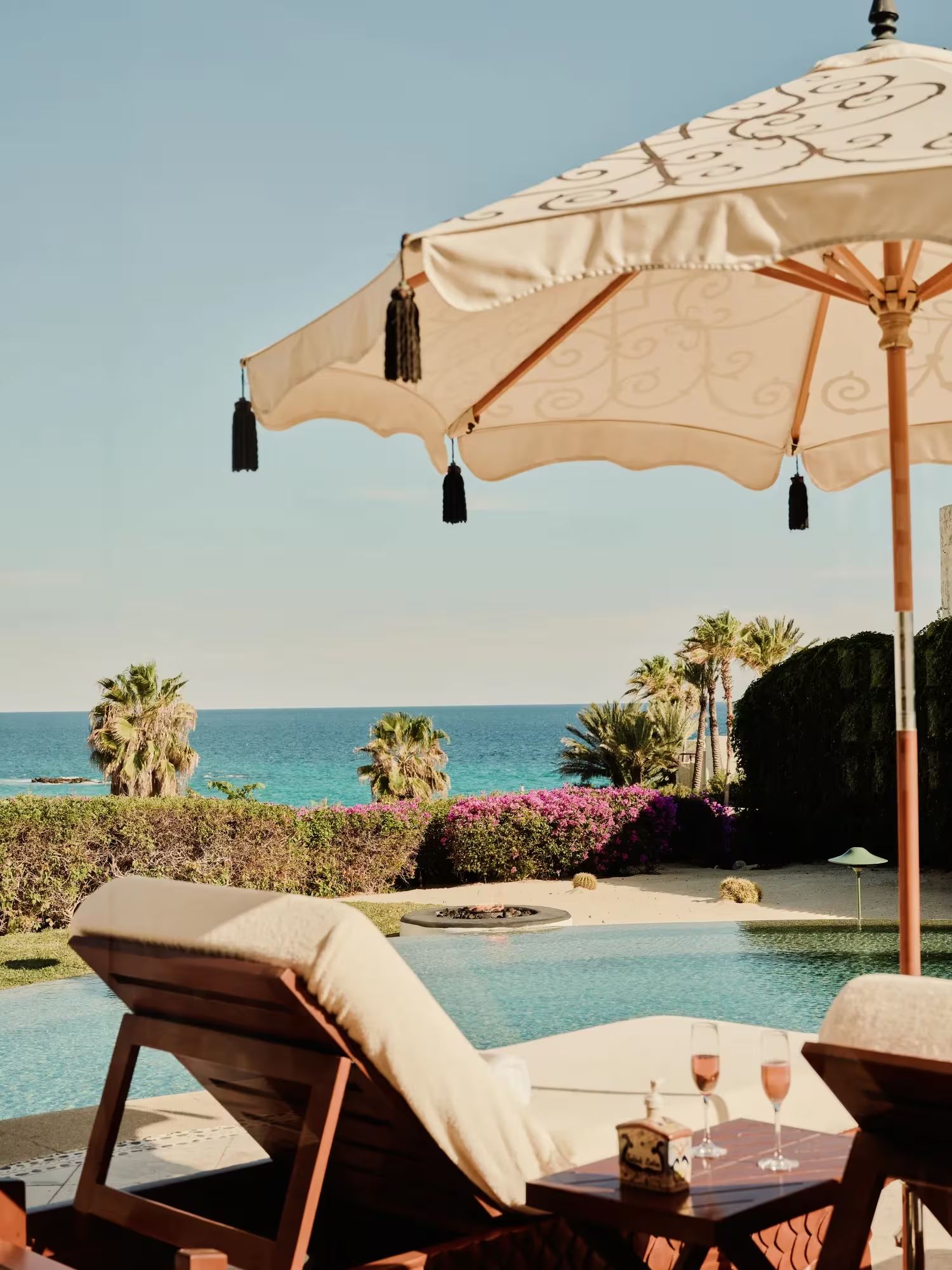 Loungers under a tasselled umbrella beside a pool, with palms and the sea beyond